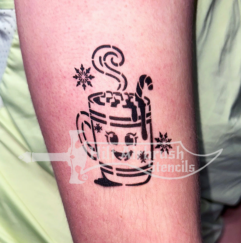 An airbrush tattoo of a hot chocolate cup with candy canes and snowflakes on a person's arm, created with a Hilt Airbrush Stencil.